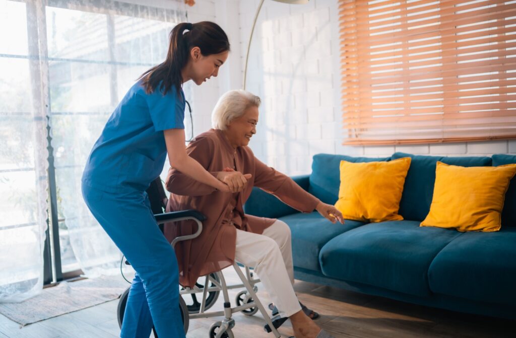 In an assisted living home, a nurse helps a senior out of their wheelchair and onto a blue couch