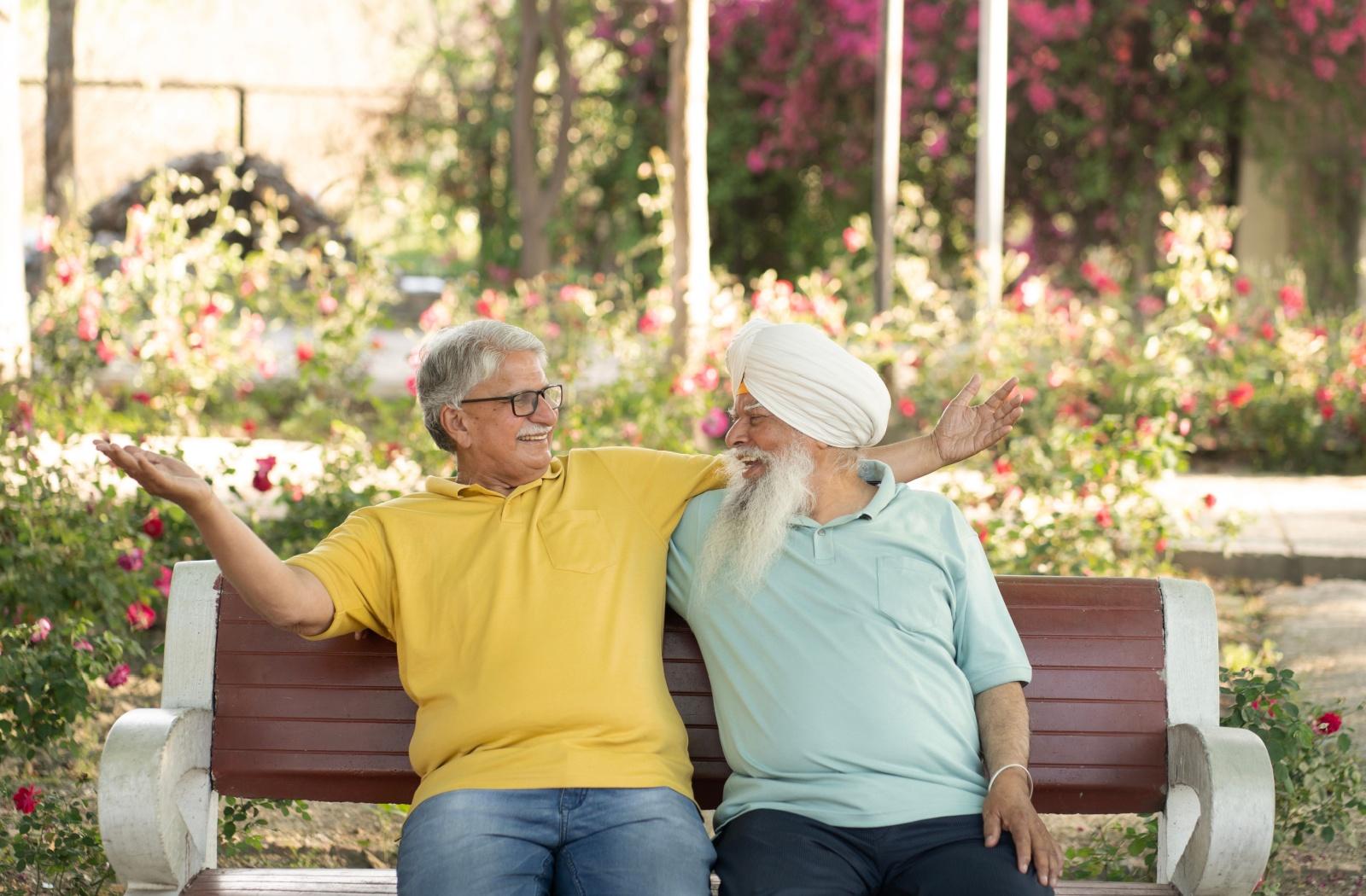 Two senior friends smile at each other while sitting on a bench in a flower garden.