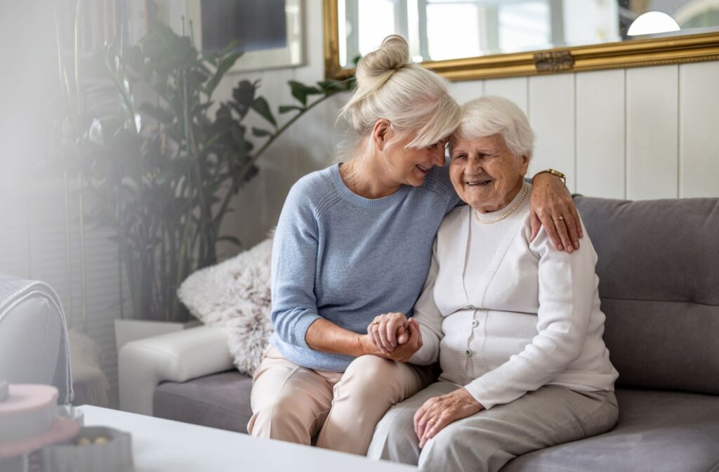An older adult embraces their senior parent while they sit on a gray sofa in an apartment.