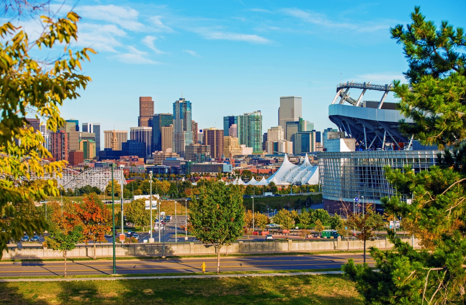 City skyline with tall buildings and a large stadium on the right, framed by trees and a clear blue sky.