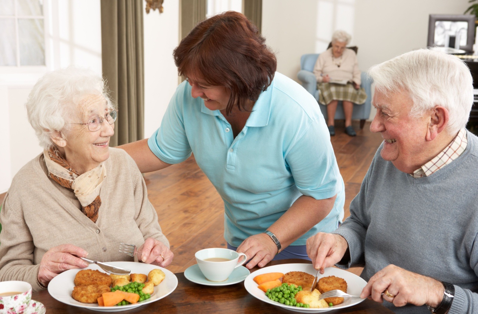 Caregiver chats with an elderly woman and man during a shared meal at a dining table.