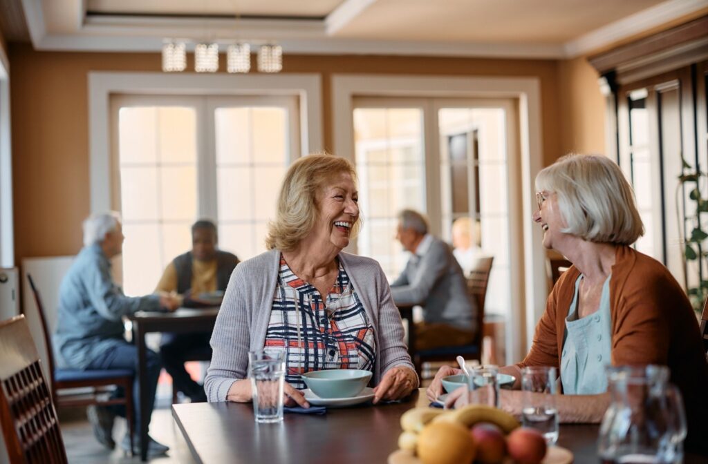 Two senior friends smile as they enjoy a healthy meal together at a senior living community.