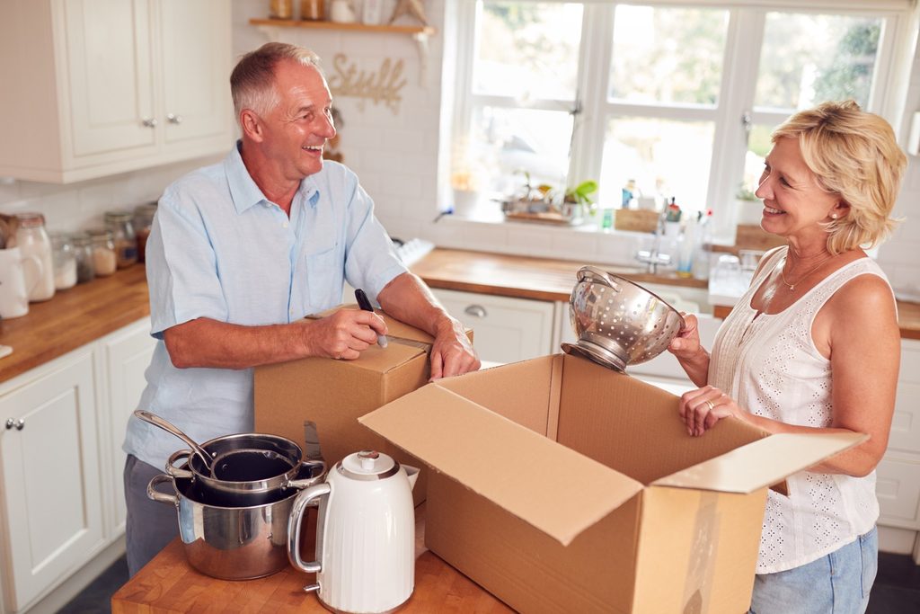 Older couple packing a box in a bright kitchen, smiling as they load cookware into a cardboard box to move home.