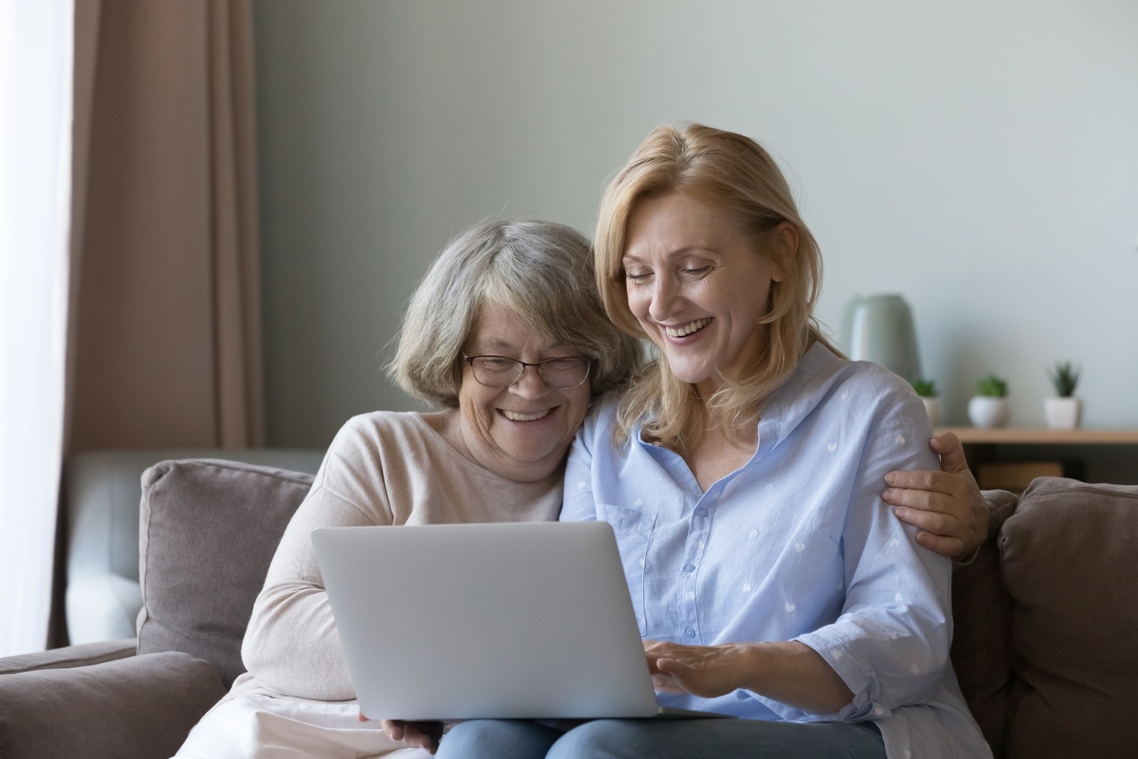 Adult daughter and old mother sit on sofa with laptop choosing assisted living via internet, web surfing together at home.