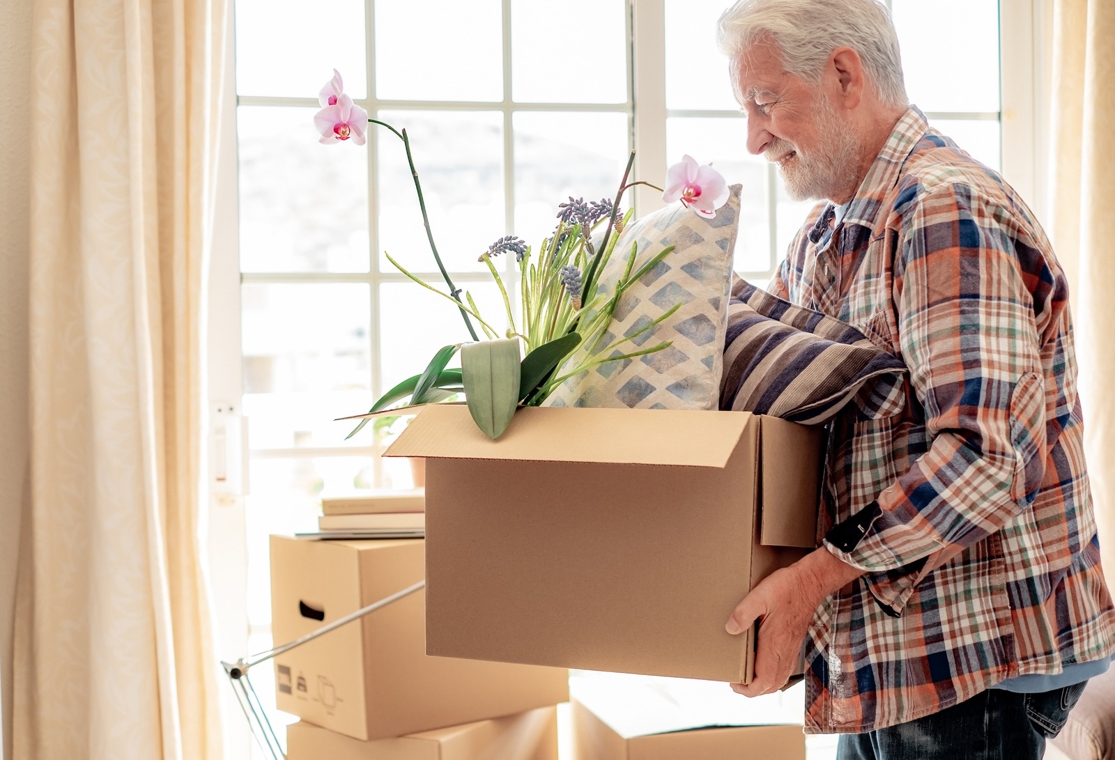 Older man packing a cardboard box with a potted orchid and striped pillow in a bright room